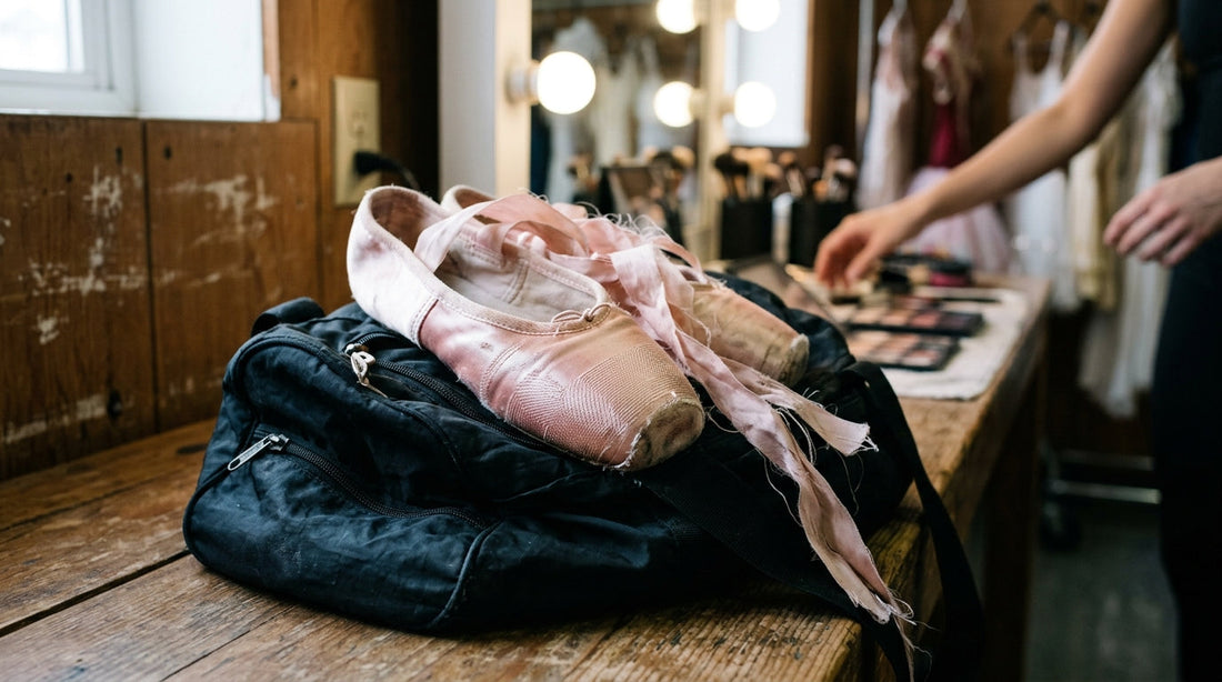 A pair of pink satin pointe shoes with messy ribbons resting on a black unzipped dance bag on a wooden dressing room bench.