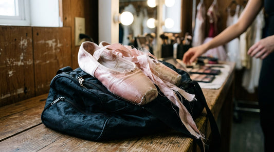 A pair of pink satin pointe shoes with messy ribbons resting on a black unzipped dance bag on a wooden dressing room bench.