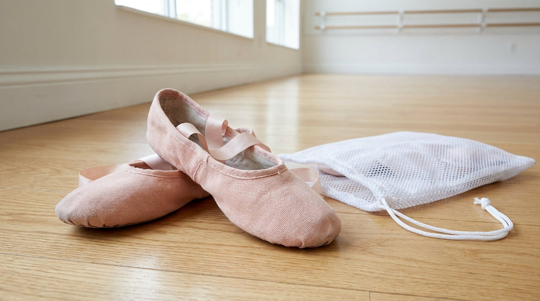A pair of pink canvas ballet slippers with satin ribbons resting on a light oak dance floor next to a white mesh laundry bag.