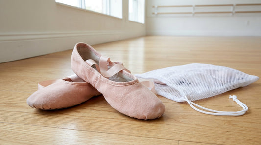 A pair of pink canvas ballet slippers with satin ribbons resting on a light oak dance floor next to a white mesh laundry bag.