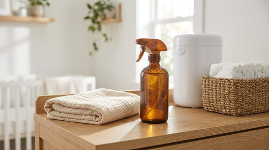 An amber glass spray bottle and a white diaper pail in a sunlit nursery with a muslin blanket.