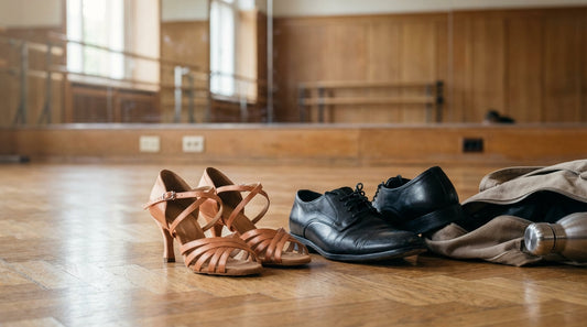 Elegant satin Latin heels and men's leather ballroom shoes resting on a polished parquet dance floor next to an unzipped gear bag.