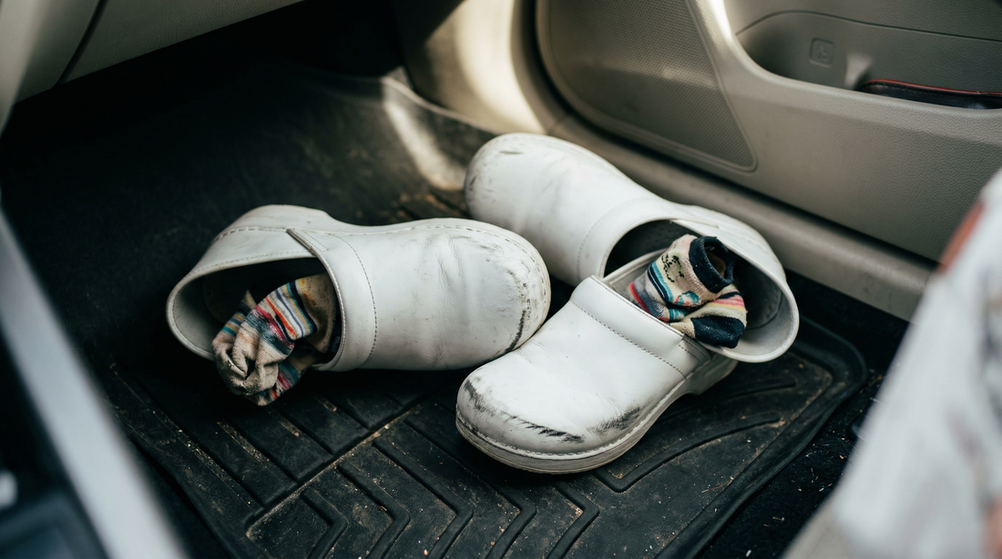 White nursing clogs and patterned compression socks kicked off on a black car floor mat, showing smelly nursing shoes after a shift.