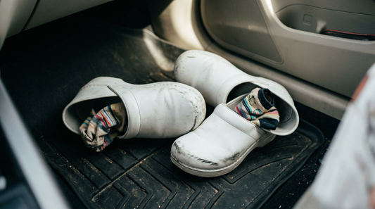 White nursing clogs and patterned compression socks kicked off on a black car floor mat, showing smelly nursing shoes after a shift.