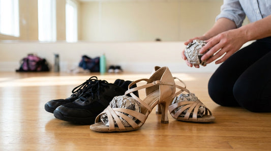 Strappy satin salsa heels and dance sneakers with crumpled newspaper stuffed inside on a polished wood floor.