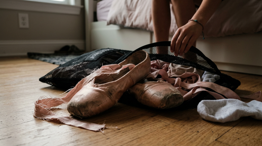 Worn pink satin ballet slippers with frayed ribbons spilling out of an unzipped dance bag on a wooden floor.