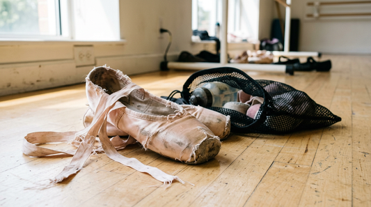 Worn pink satin pointe shoes with frayed ribbons and a mesh bag on a dance studio floor showing signs of pointe shoe odor damage.