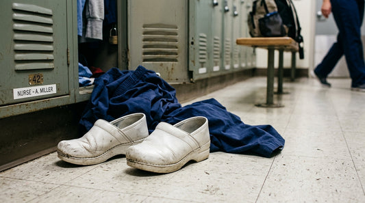 White leather nursing clogs on a locker room floor next to navy blue scrubs and a metal locker.