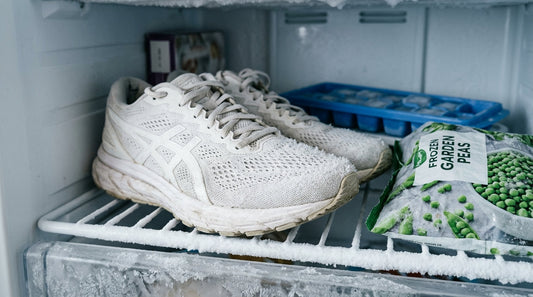 A pair of white mesh running shoes sitting on a freezer shelf next to a bag of frozen peas and a plastic ice cube tray.
